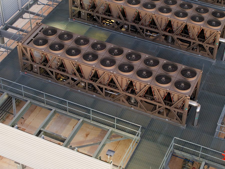 Aerial drone photograph of a large flat-roofed industrial building in the desert with a row of grey cooling-tower units on the roof and a paved lot around it.