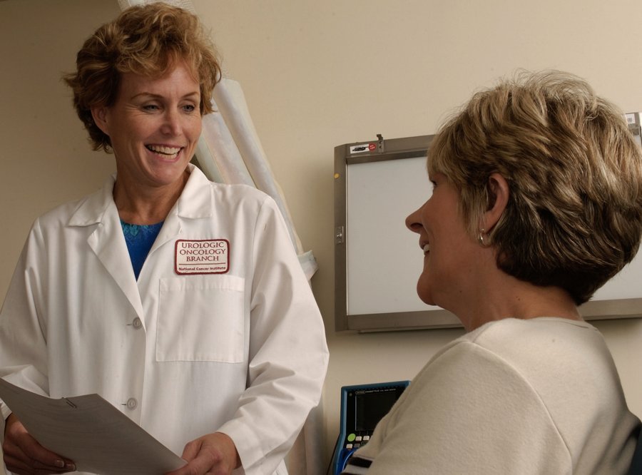 A physician in a white coat sits across a desk from a patient, both facing each other in conversation, with paperwork between them.