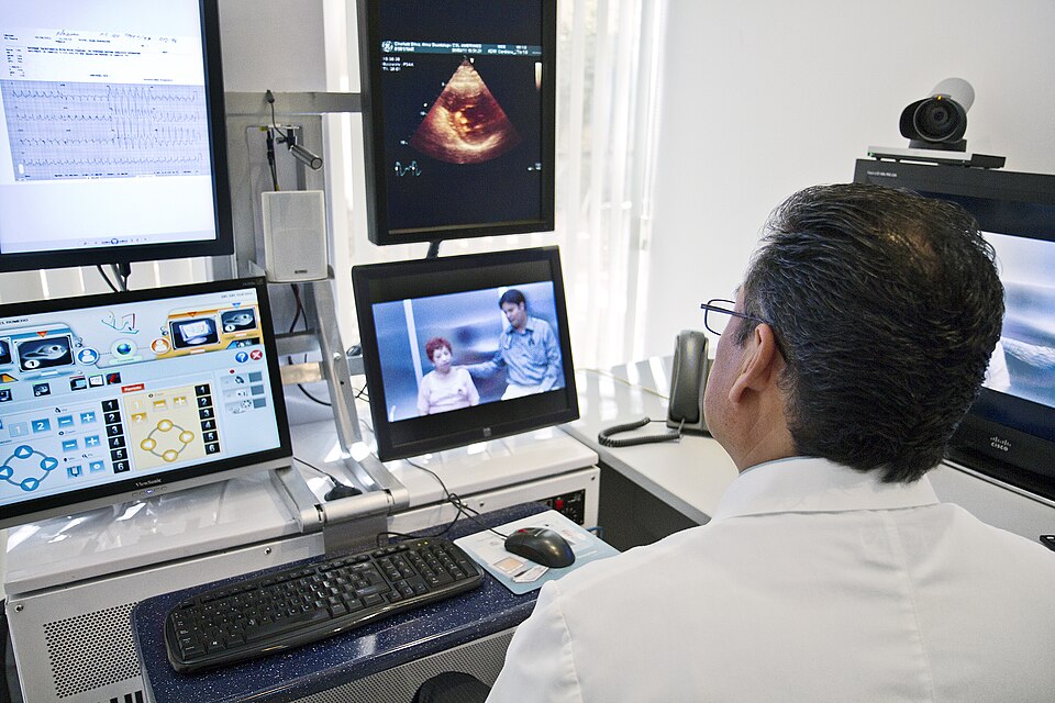 A physician in a white coat seen from behind, facing four monitors showing an ECG, an echocardiogram, and a video call with two people.